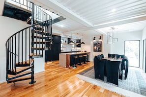 A modern kitchen with a central island, a dining area with a wooden table and chairs, and a spiral staircase with black railings.