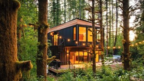A modern cabin with a glass facade, surrounded by forest. It features a deck with outdoor seating and string lights. - Pacific Bin - Sauna / Hot Tub / Steam Room (Monroe)