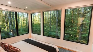 A bedroom with a large bed, a window with a forest view, and a small rug on the floor. - Pacific Bin - Sauna / Hot Tub / Steam Room (Monroe)