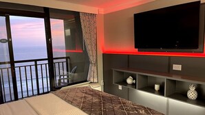 A bedroom with a bed, a TV mounted on the wall, and a balcony with a view of the sea.
