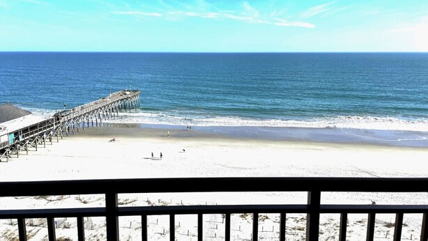 A beach with a pier extending into the ocean, a clear sky, and a view of the beach from a balcony.