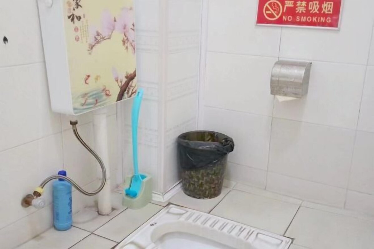 A restroom with a toilet, a hand dryer, a trash bin, and a no-smoking sign.