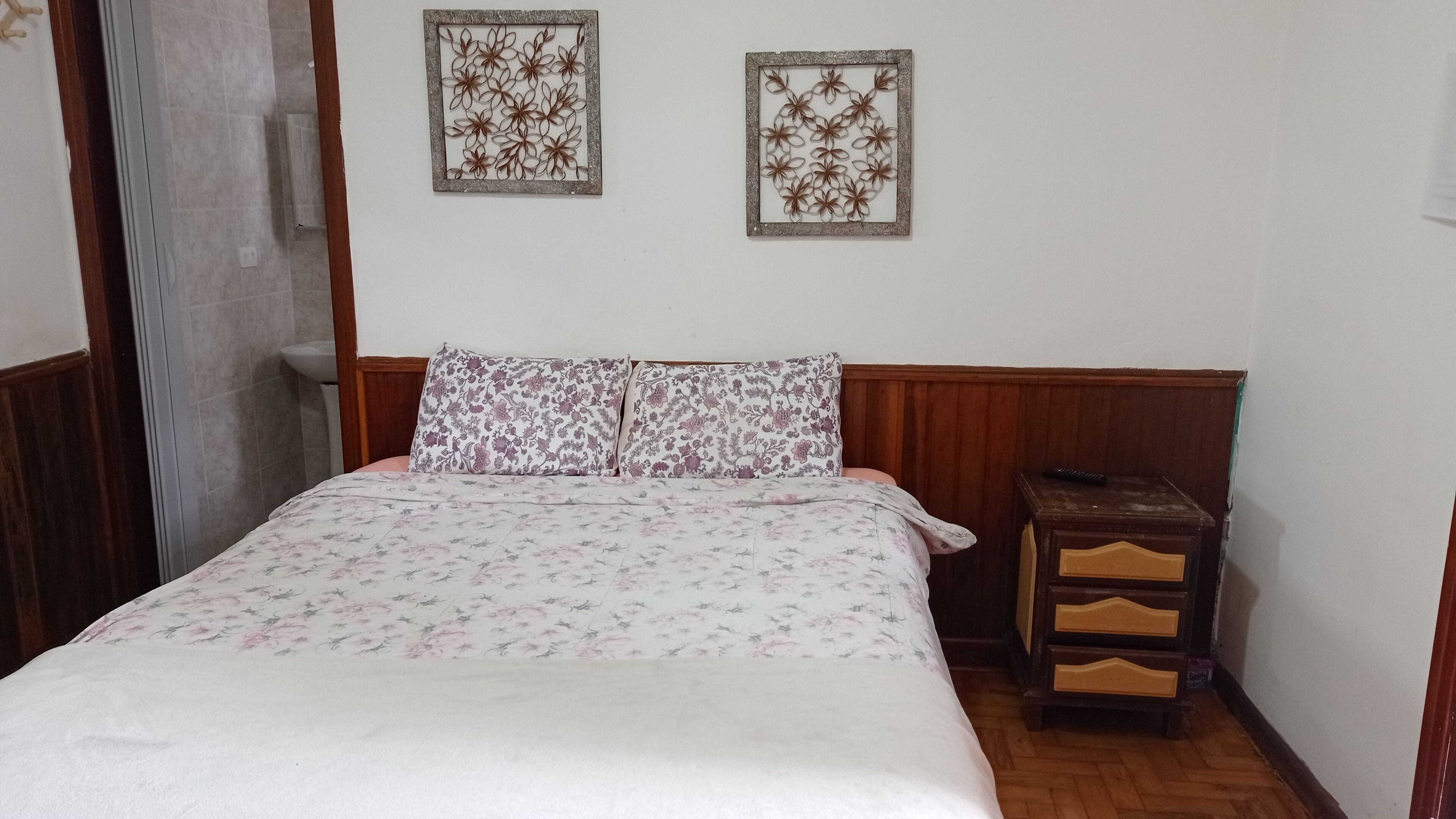 A bedroom with a bed, floral-patterned bedding, two wall-mounted decorative panels, a wooden nightstand with drawers, and a wooden headboard.