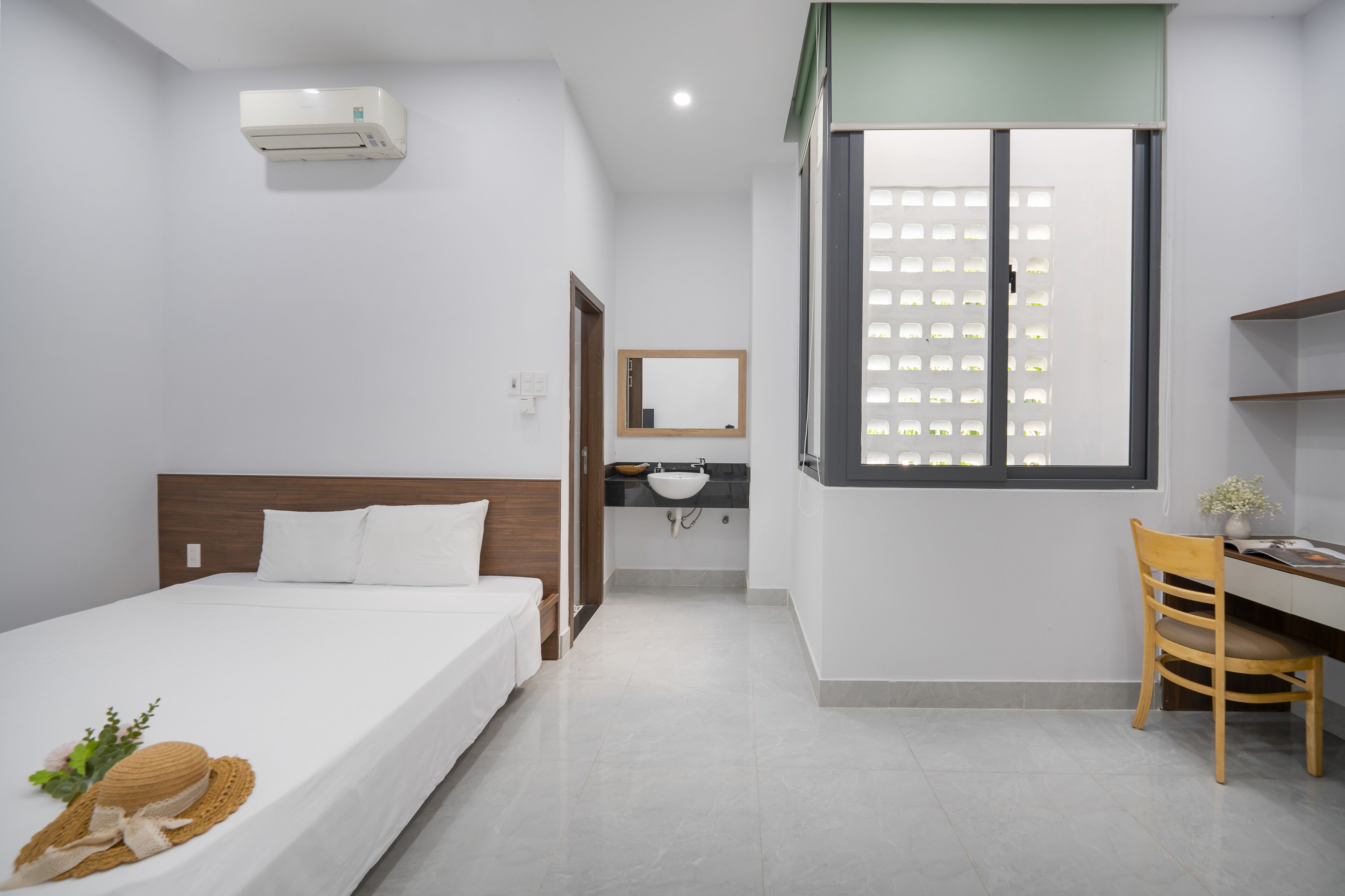 Een moderne hotelkamer met een bed, een bureau met stoel, een raam met zonwering, een badkamer met wastafel, en een airconditioning.