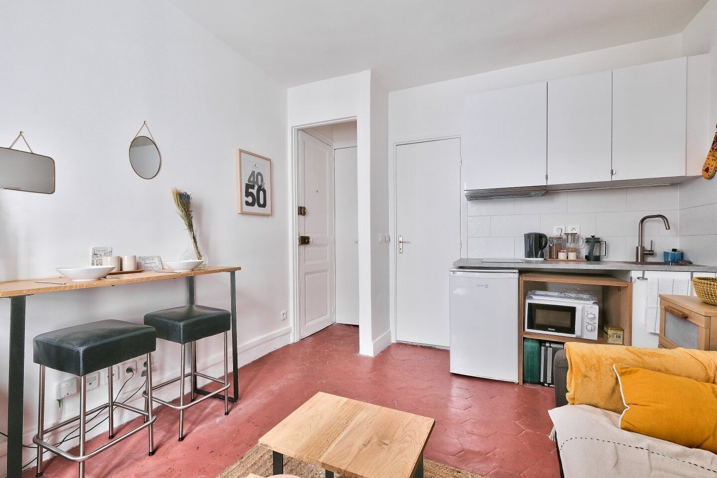 Une cuisine compacte avec des meubles blancs, un four à micro-ondes et un évier. On y trouve également un petit coin repas avec deux tabourets et une table.