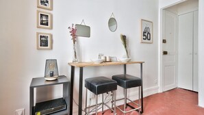 A modern interior with a wooden table, two black stools, a small side table with a metal basket, a mirror, and framed pictures on the wall.