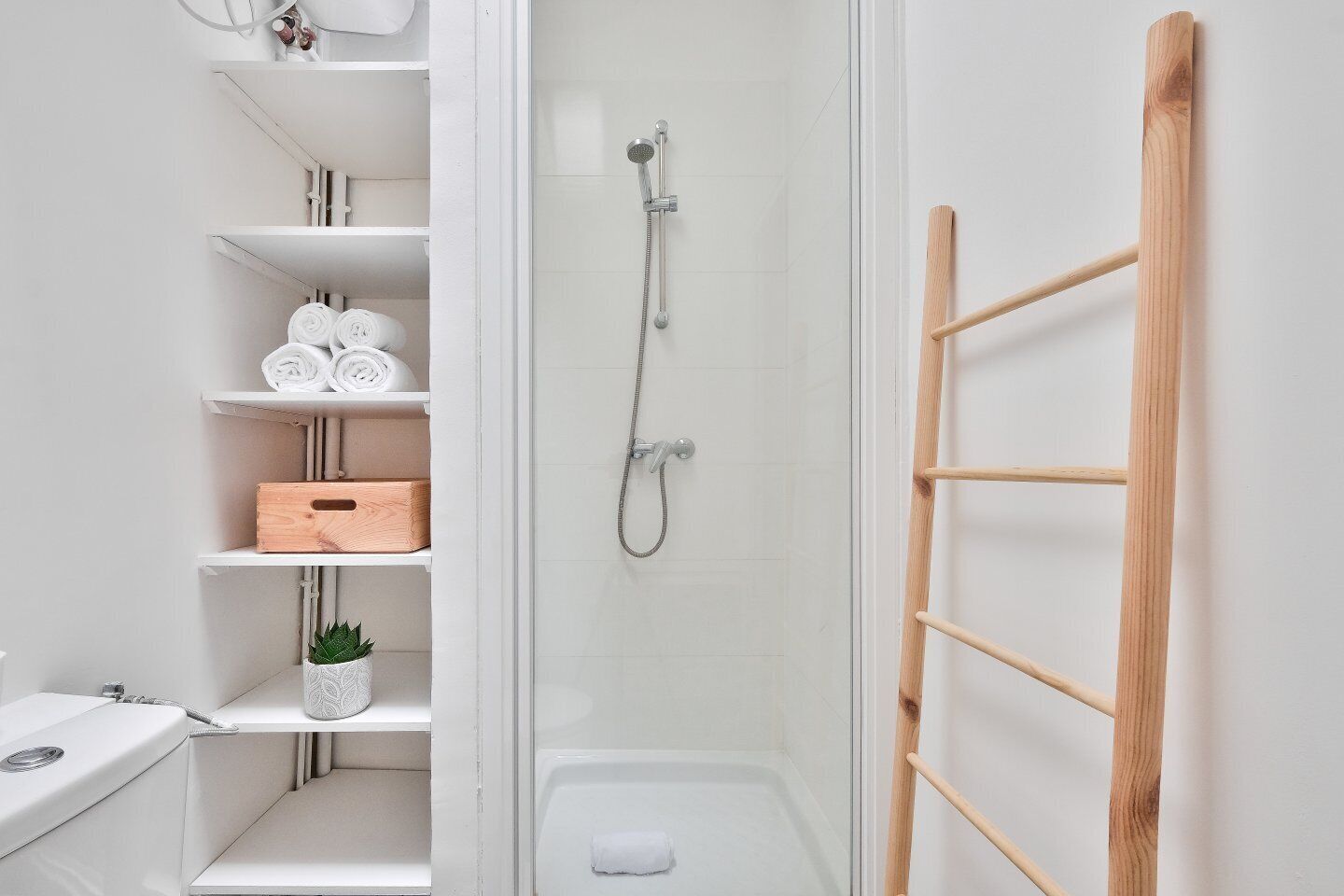 A modern bathroom with a glass shower enclosure, white shelves with towels and toiletries, and a wooden ladder against the wall.
