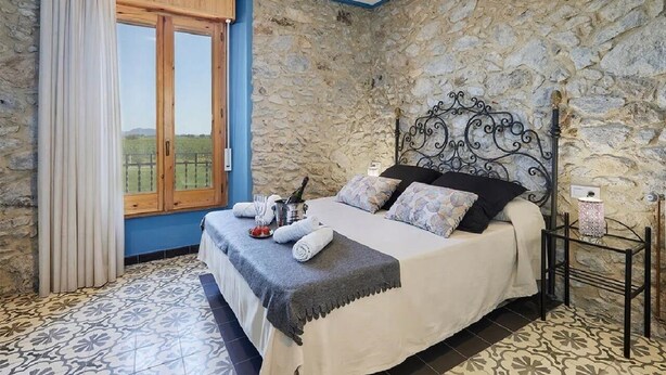 A bedroom with a stone wall, a bed with a metal headboard, a window with curtains, and a patterned floor. - Mas Ribas Turismo Rural (Ventallo)