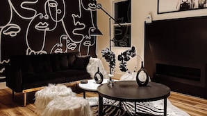 Ein modernes Wohnzimmer mit einem schwarzen Sofa, einem runden Couchtisch, einem weißen, flauschigen Hocker, einem Teppich mit Zebra-Muster und einer Wand mit abstrakter Kunst.
