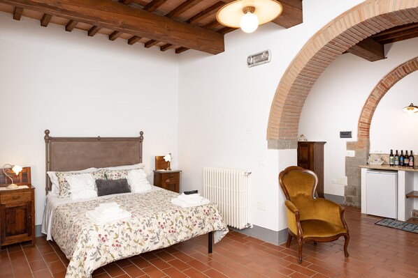 Un dormitorio con cama, mesita de noche, silla, radiador y una pequeña cocina con mostrador y botellas.