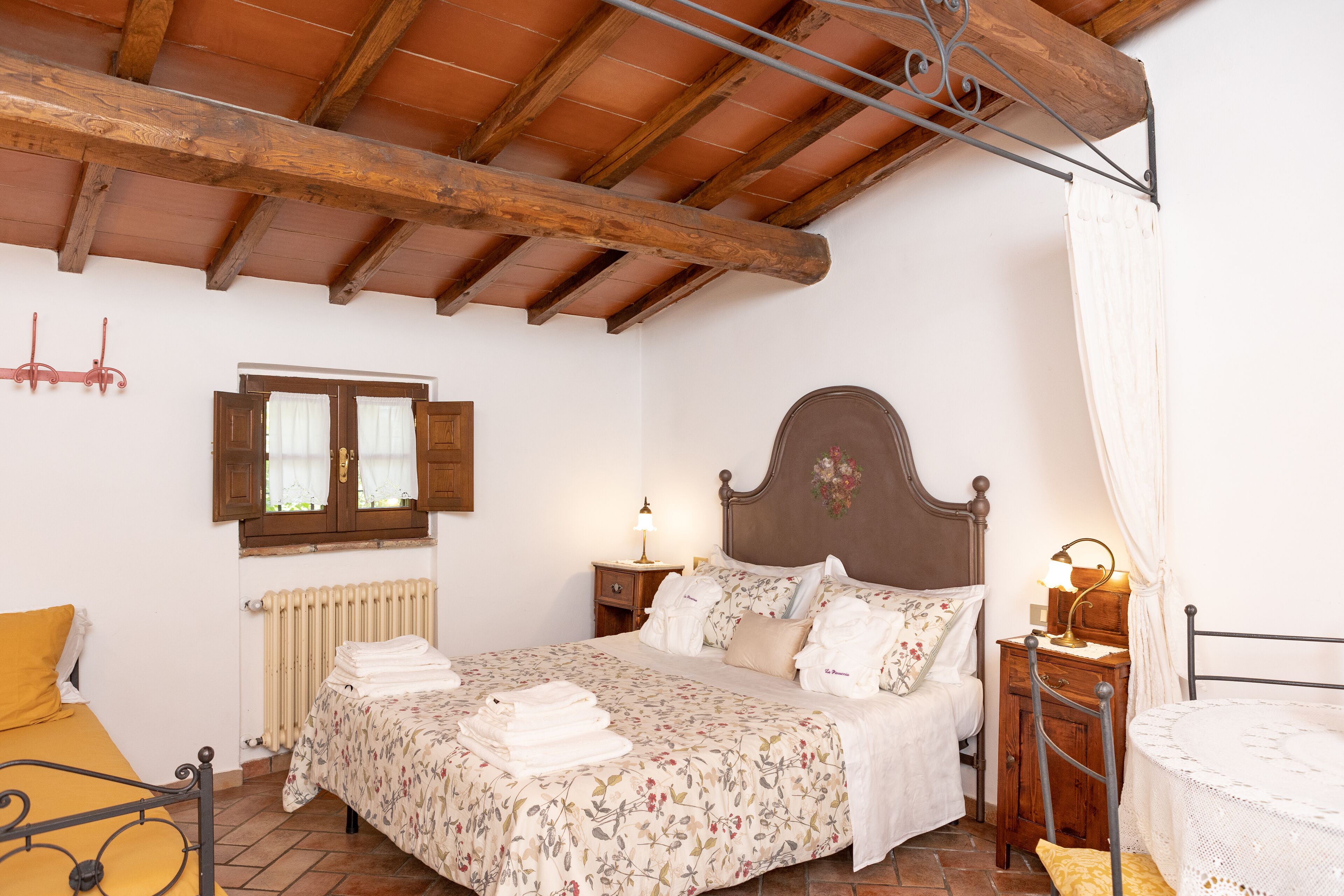 Un dormitorio con techo de madera, cama con ropa de cama floral, mesita de noche con lámpara y una silla con una mesa.