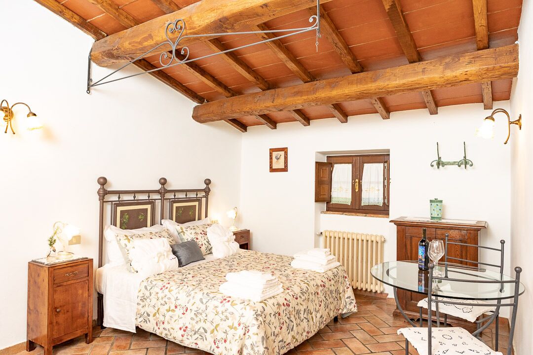 A bedroom with a wooden ceiling, a bed with floral bedding, a bedside table with a lamp, a small cabinet, and a glass table with a chair.