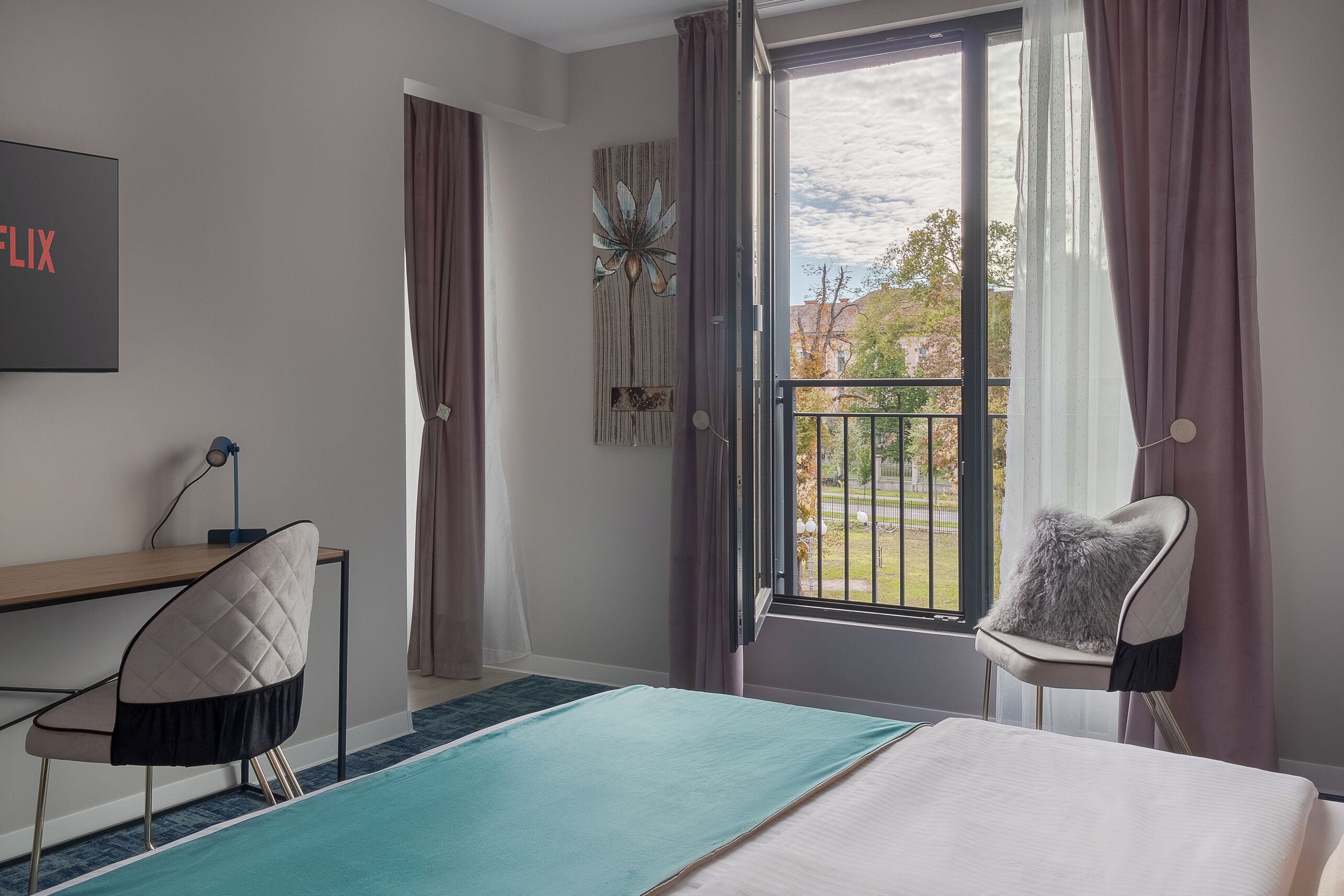 A hotel room with a bed, a desk, a chair, a large window with curtains, and a view of the outside.