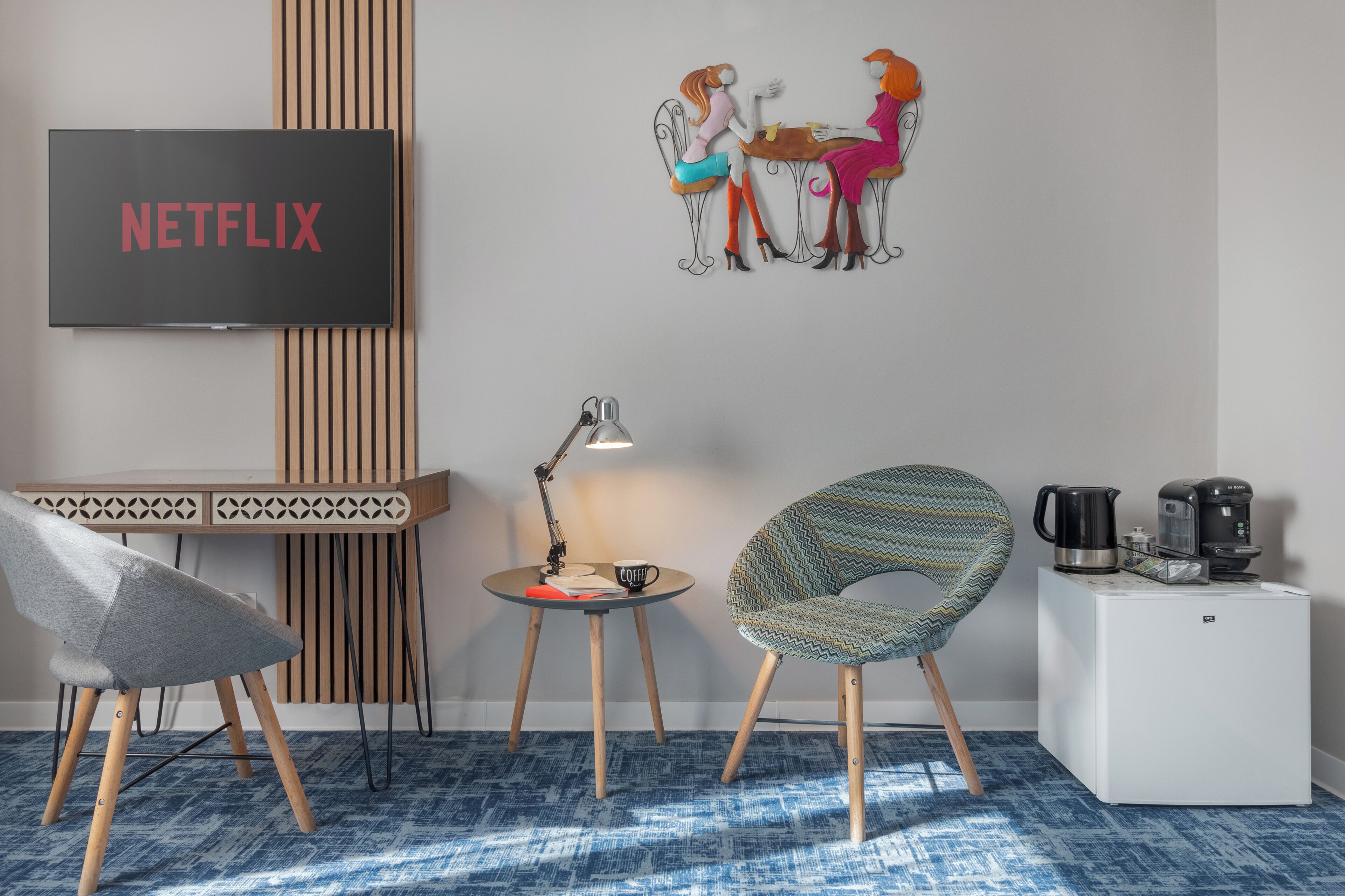 A modern room with a flat-screen TV displaying 'NETFLIX', a small round table with a lamp, a patterned armchair, a coffee station, and a wall-mounted artwork of two women.