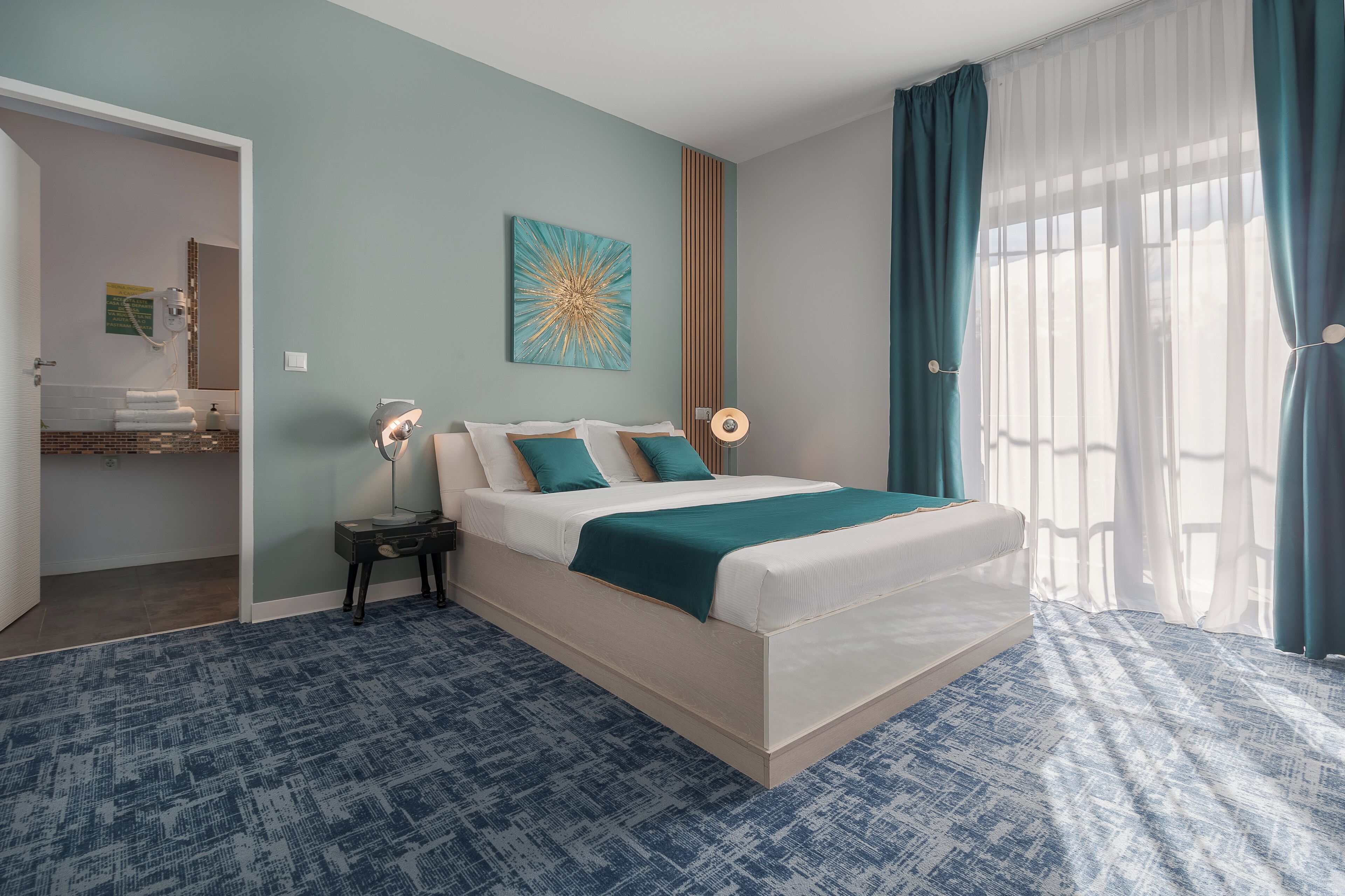 A bedroom with a bed, a bedside table, a wall-mounted lamp, a painting, and a window with curtains.