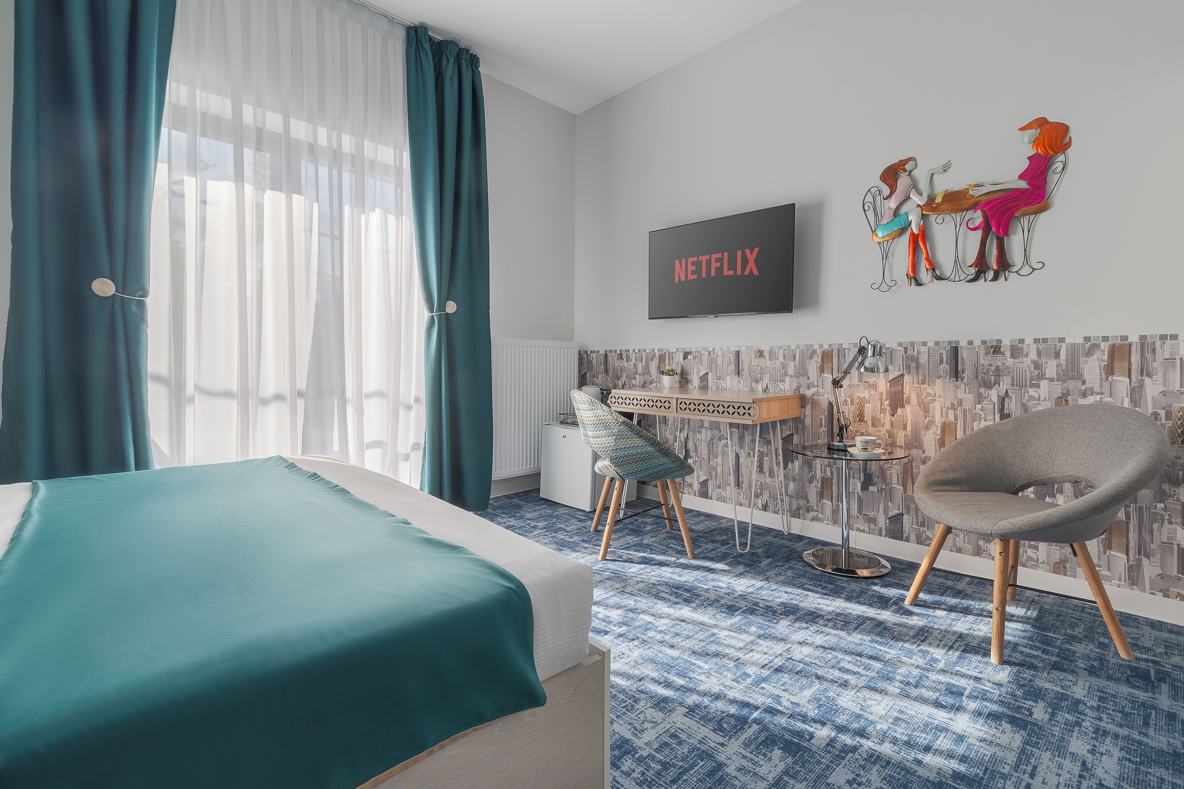 Quarto de hotel com cama, escrivaninha, cadeira, TV na parede e papel de parede com vista urbana.