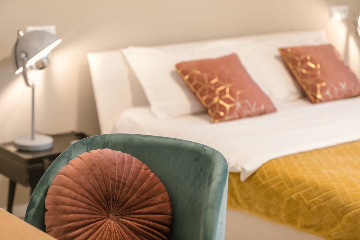 A bed with white linens and two decorative pillows, a teal armchair with a brown cushion, and a bedside table with a lamp.