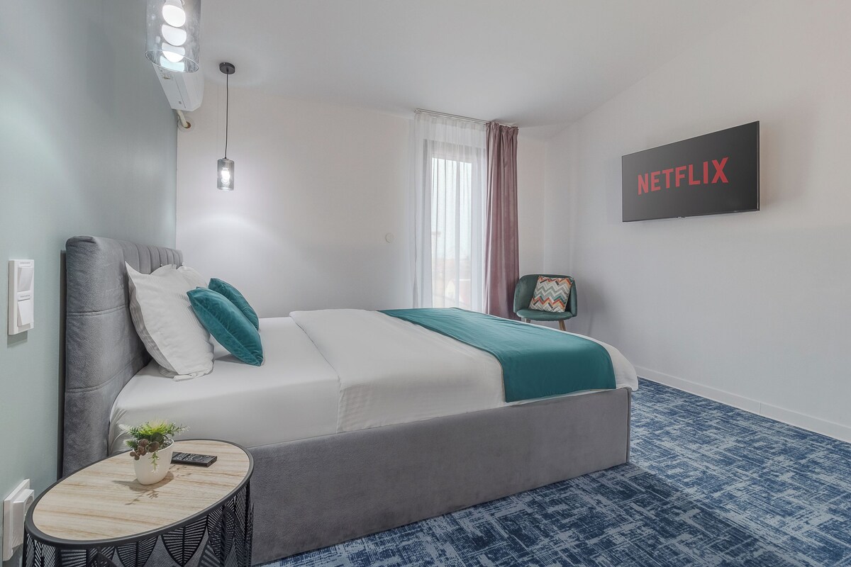 A bedroom with a bed, a bedside table with a plant, a wall-mounted TV, and a window with curtains.