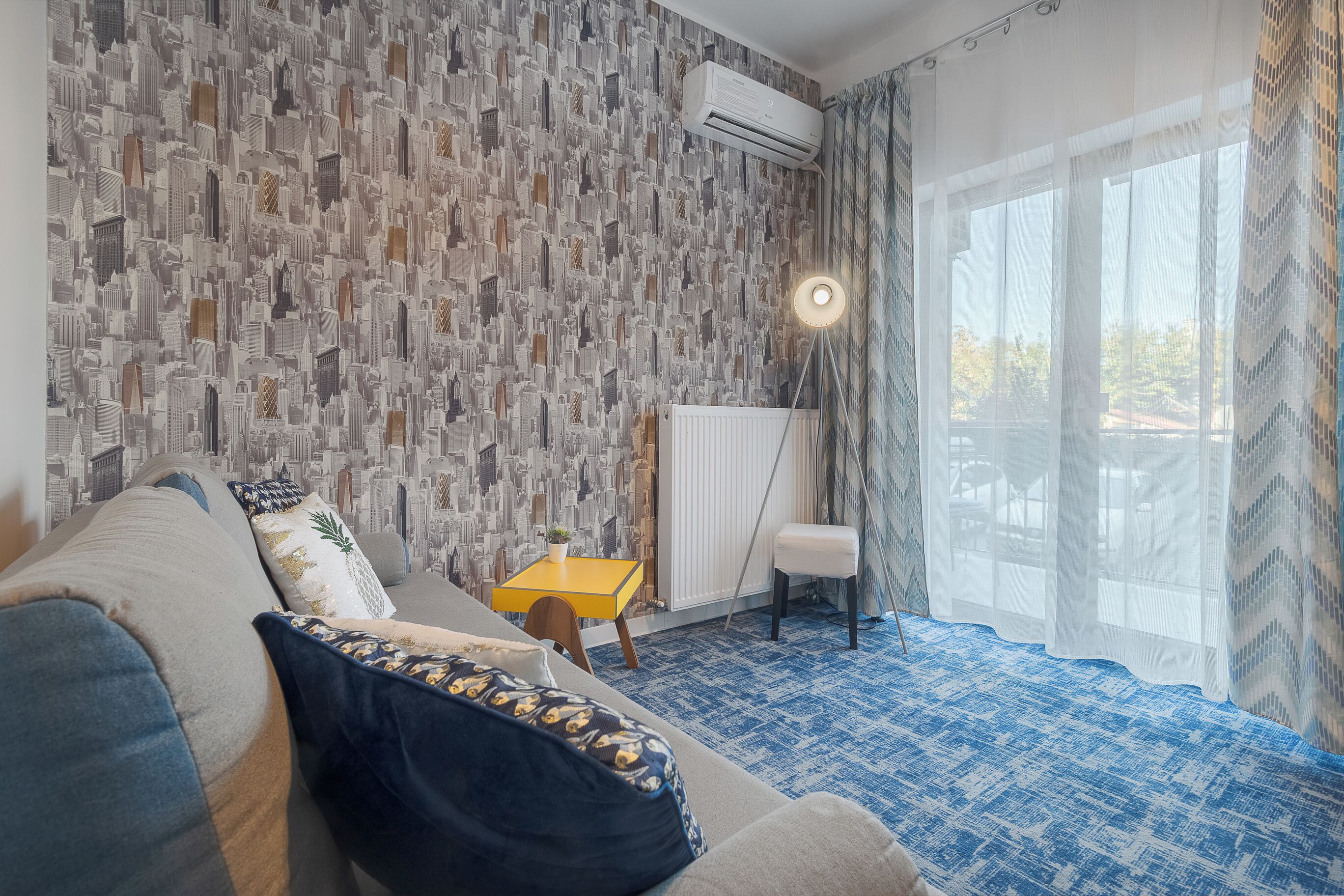 A room with a patterned cityscape wallpaper, a sofa with patterned cushions, a small yellow table, a chair, and a large window with sheer curtains.