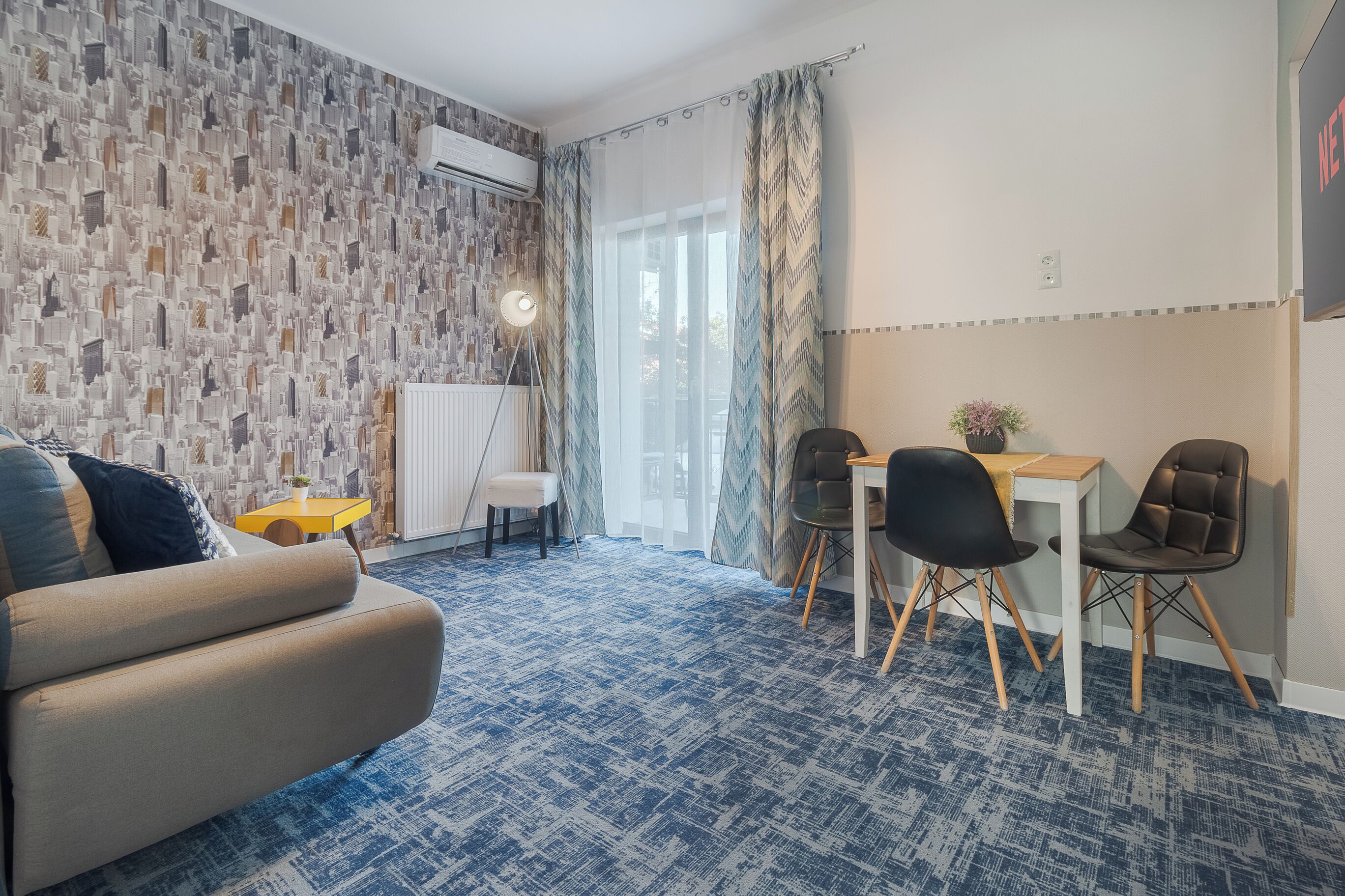 Una habitación moderna con un papel tapiz estampado, alfombra azul, sofá, mesa con dos sillas y ventana con cortinas.