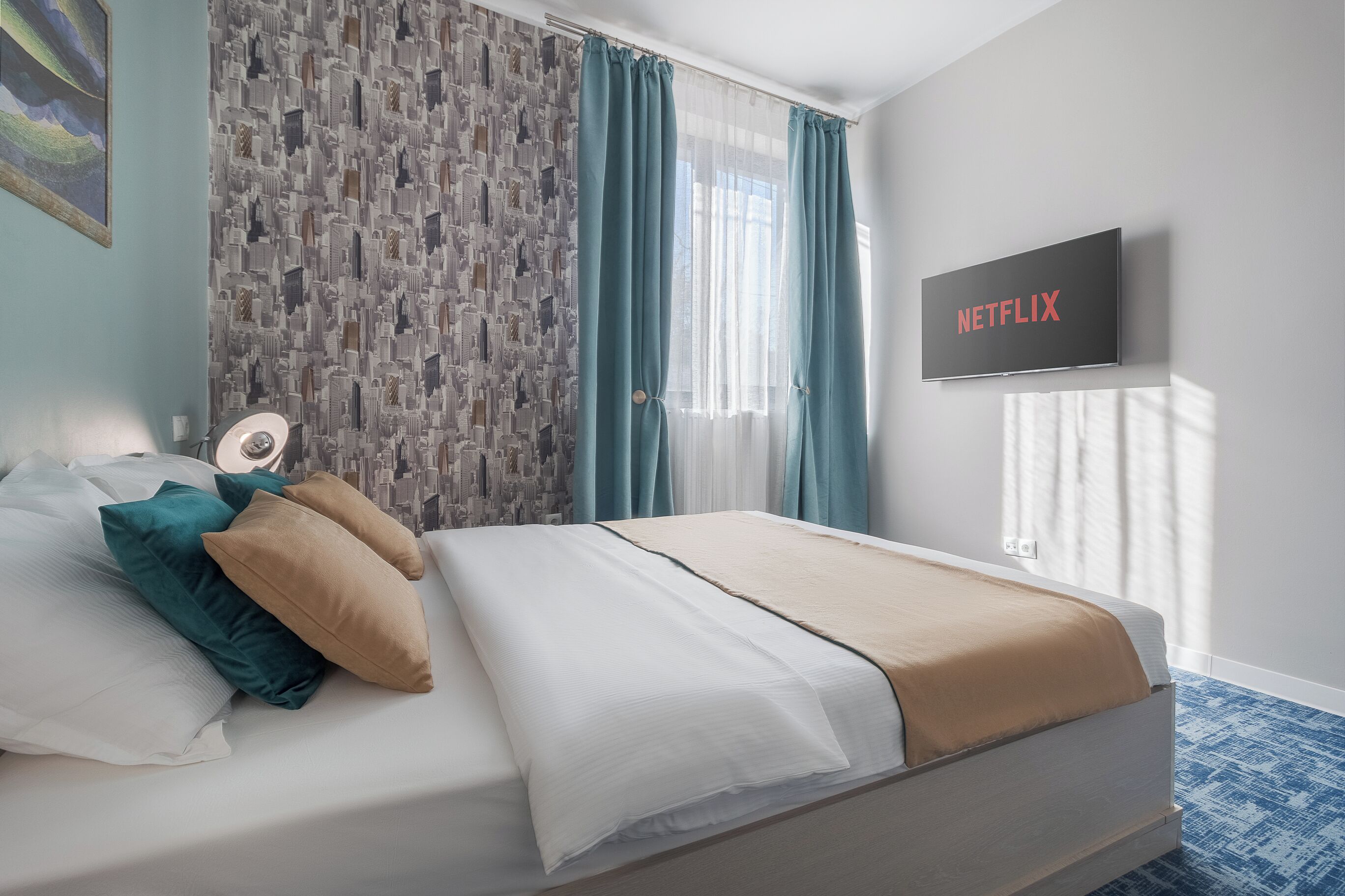 Una cama bien hecha con edredón blanco y beige, almohadas verde azulado y beige, y una alfombra estampada azul. Un televisor en la pared muestra el logo de Netflix.