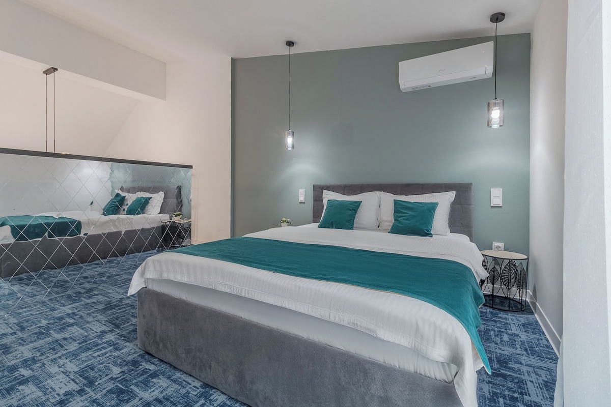 A modern hotel room with a large bed, a bedside table, a small round table, and a wall-mounted air conditioner.