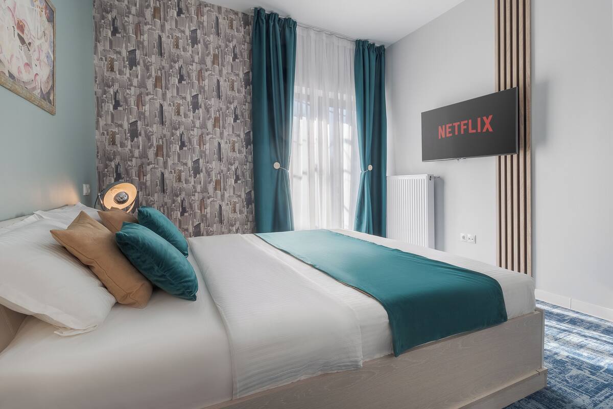 A modern bedroom with a bed, teal curtains, a wall-mounted TV displaying "NETFLIX," and a patterned wall.