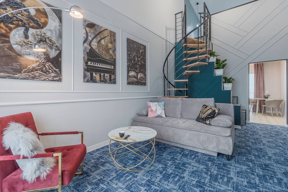 A modern living room with a grey sofa, red armchair, and a round coffee table. A spiral staircase with a teal accent leads to an upper level.
