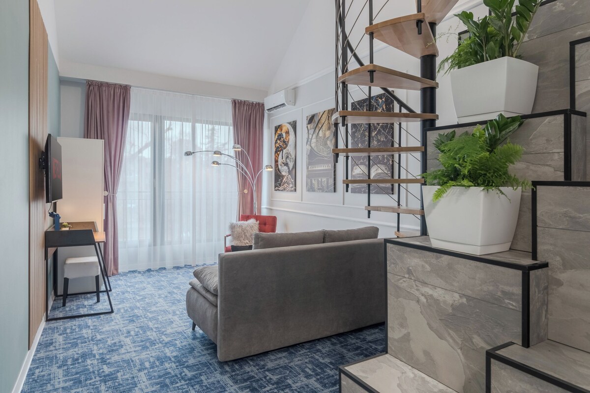 A modern living room with a grey sofa, a small table with a chair, a TV, and a staircase with plants.