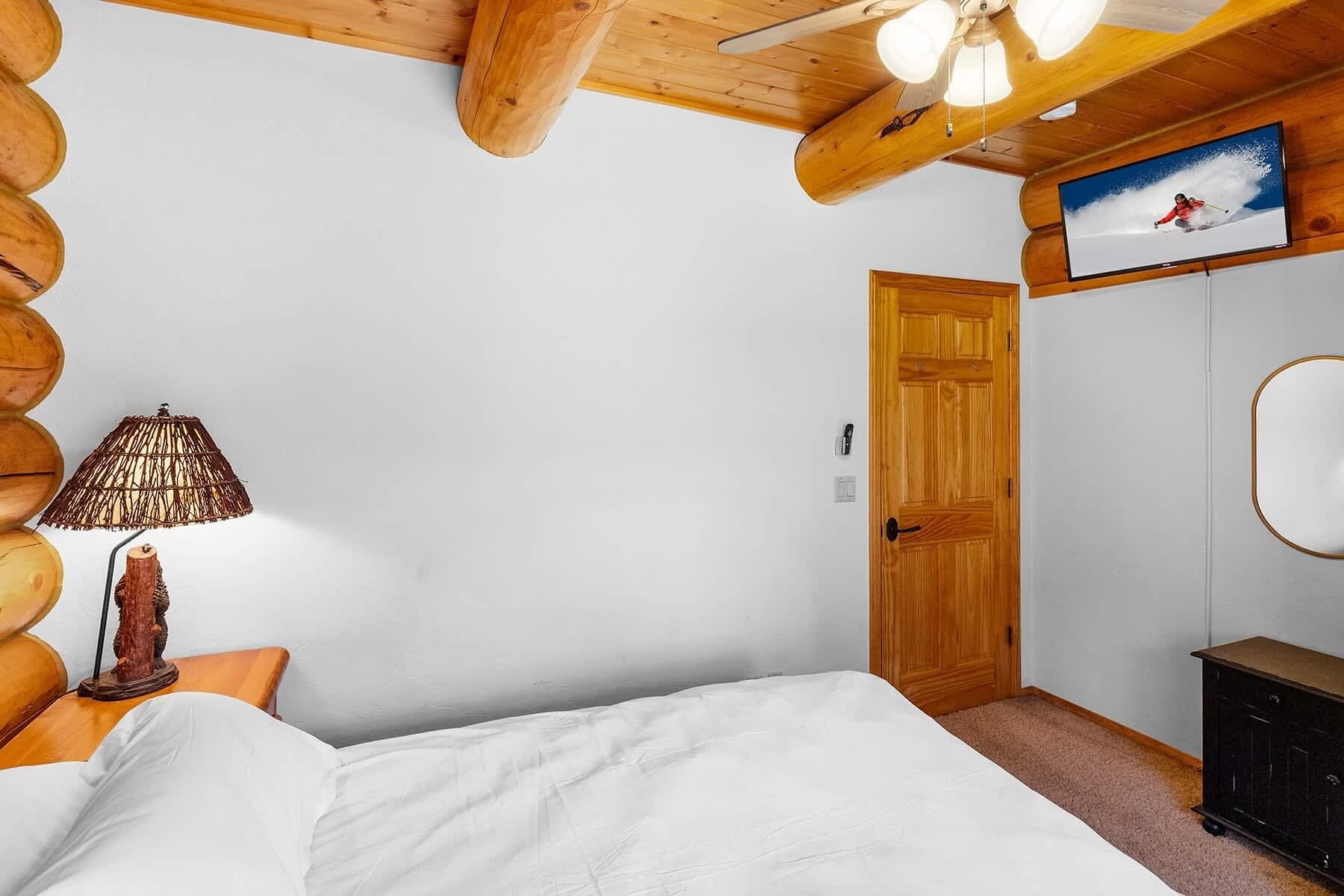 A bedroom with a wooden ceiling, a bed with white bedding, a wooden door, a lamp, and a television on the wall.