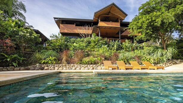 Ein modernes Haus mit Pool, umgeben von üppiger Vegetation und einer Steinmauer.