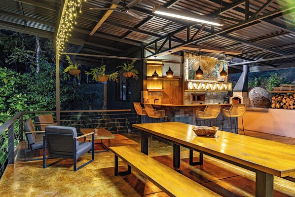 An outdoor dining area with wooden tables and chairs, hanging plants, and a stone fireplace.