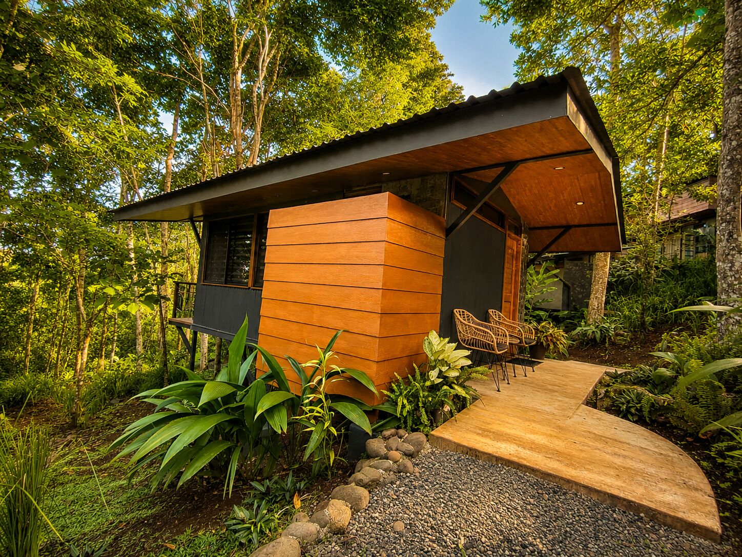 A modern wooden cabin with a curved roof, surrounded by lush greenery and a gravel path leading to a seating area with wicker chairs.