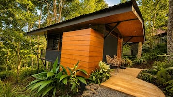 A modern wooden cabin with a curved roof, surrounded by lush greenery and a gravel path leading to a seating area with wicker chairs.