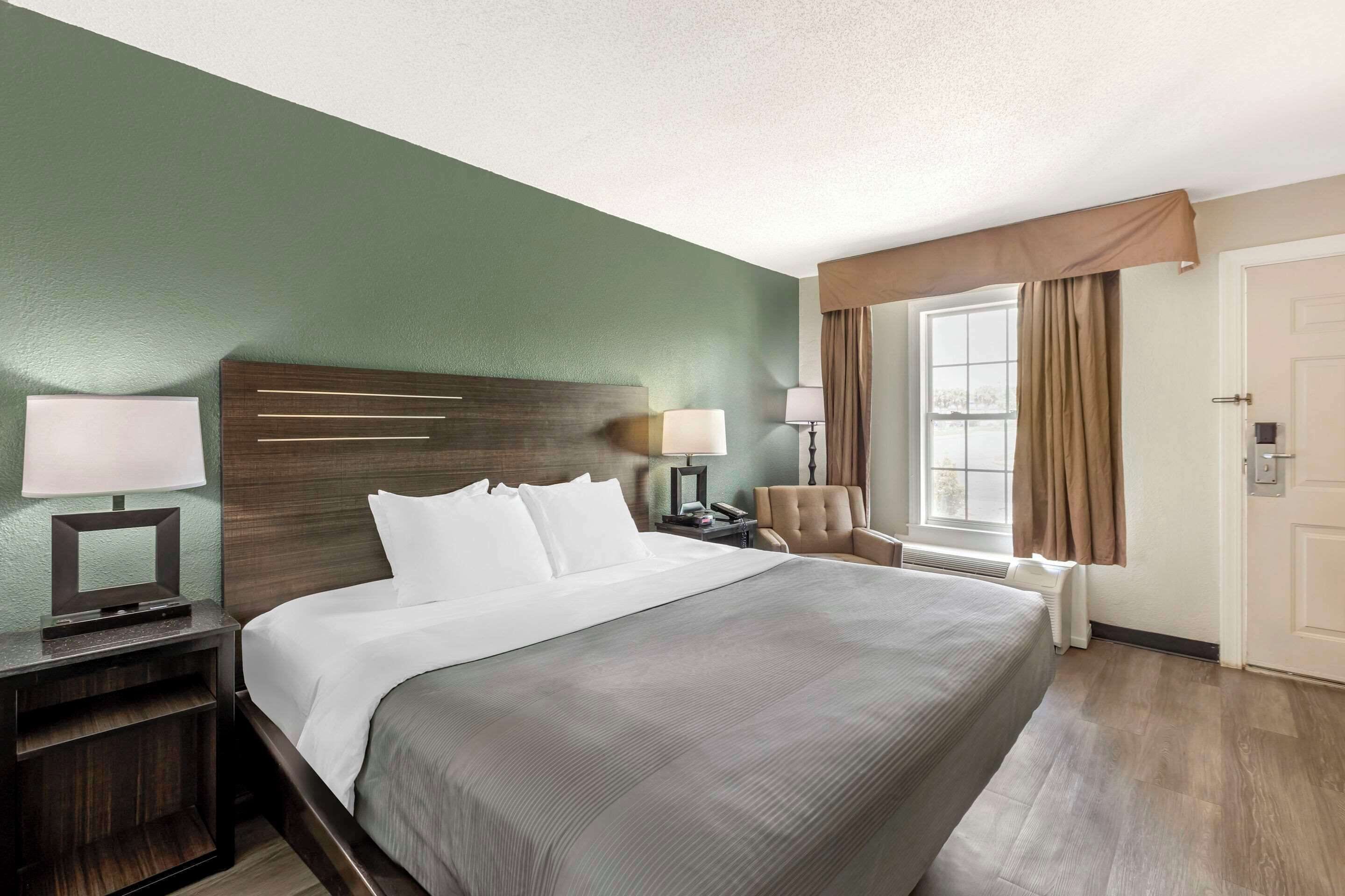 Habitación de hotel con una cama grande, una mesita de noche con lámpara, una silla y una ventana con cortinas.