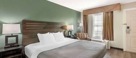 Habitación de hotel con una cama grande, una mesita de noche con lámpara, una silla y una ventana con cortinas.