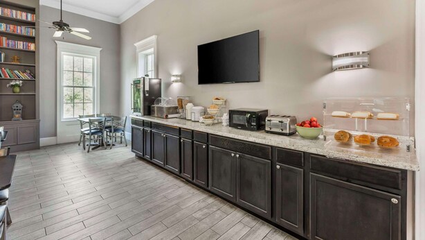 A modern kitchen with dark cabinets, a granite countertop, a ceiling fan, a flat-screen TV, and a breakfast bar with pastries. - Quality Inn & Suites Santee I-95 (Santee)
