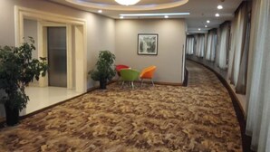 A hotel corridor with a patterned carpet, a ceiling light fixture, a framed picture on the wall, and a row of chairs along the side. - Qingyuan Hotel University of Anhui (Hefei)