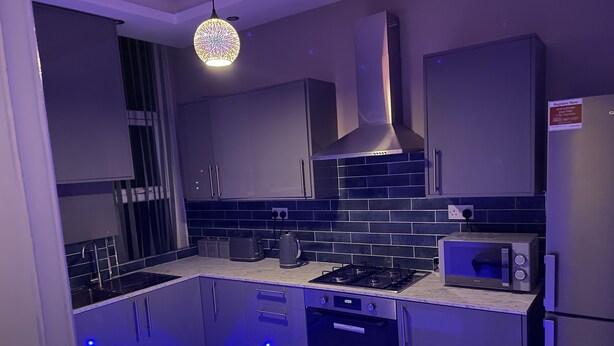 A modern kitchen with grey cabinets, a gas stove, a microwave, and blue LED strip lighting along the baseboards. - 1 Bed Flat - Sleeps 2 - Hot Tub - Parking - Wifi (Bradford)