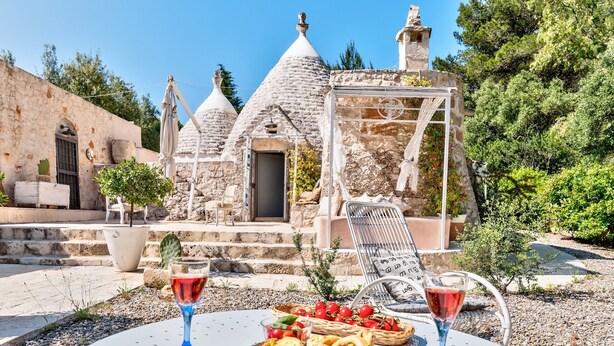 A stone building with a conical roof, a door, and a small window. There are two chairs and a table with a basket of fruit and two glasses of red wine. - Pigreco 6+2, Emma Villas (Ostuni)