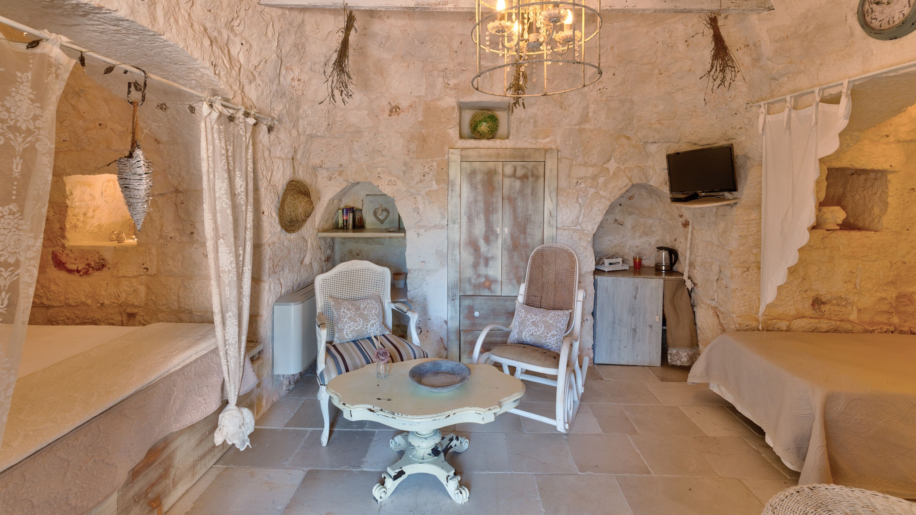 A rustic room with stone walls, a wooden door, a small table, two chairs, a television, and a chandelier.
