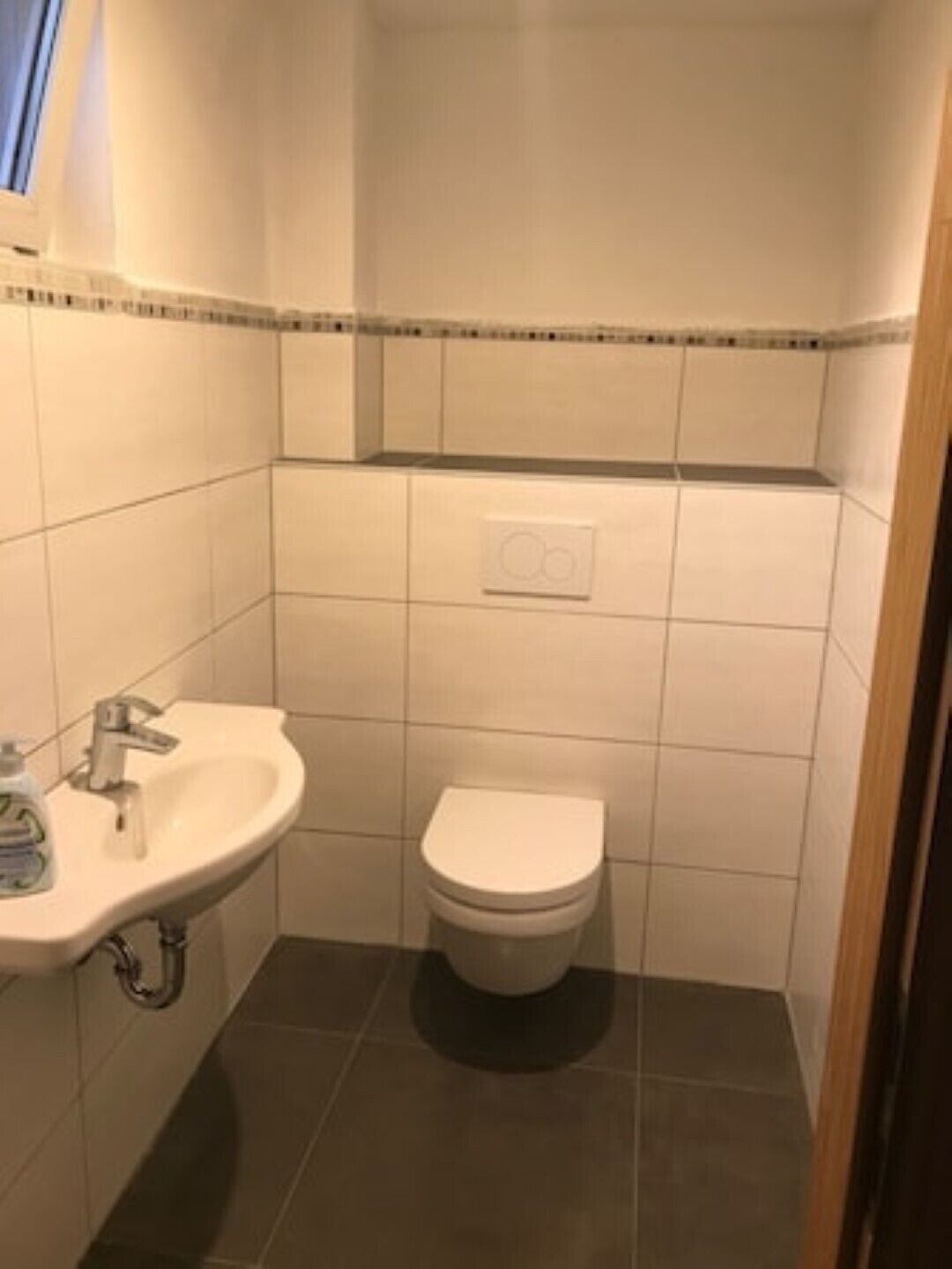 Une petite salle de bain avec un lavabo blanc, des toilettes et un interrupteur fixé au mur.