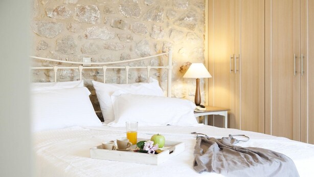 Een net opgemaakt bed met witte lakens, een dienblad met ontbijtartikelen, een stenen muur, een houten garderobe en een bedlampje.