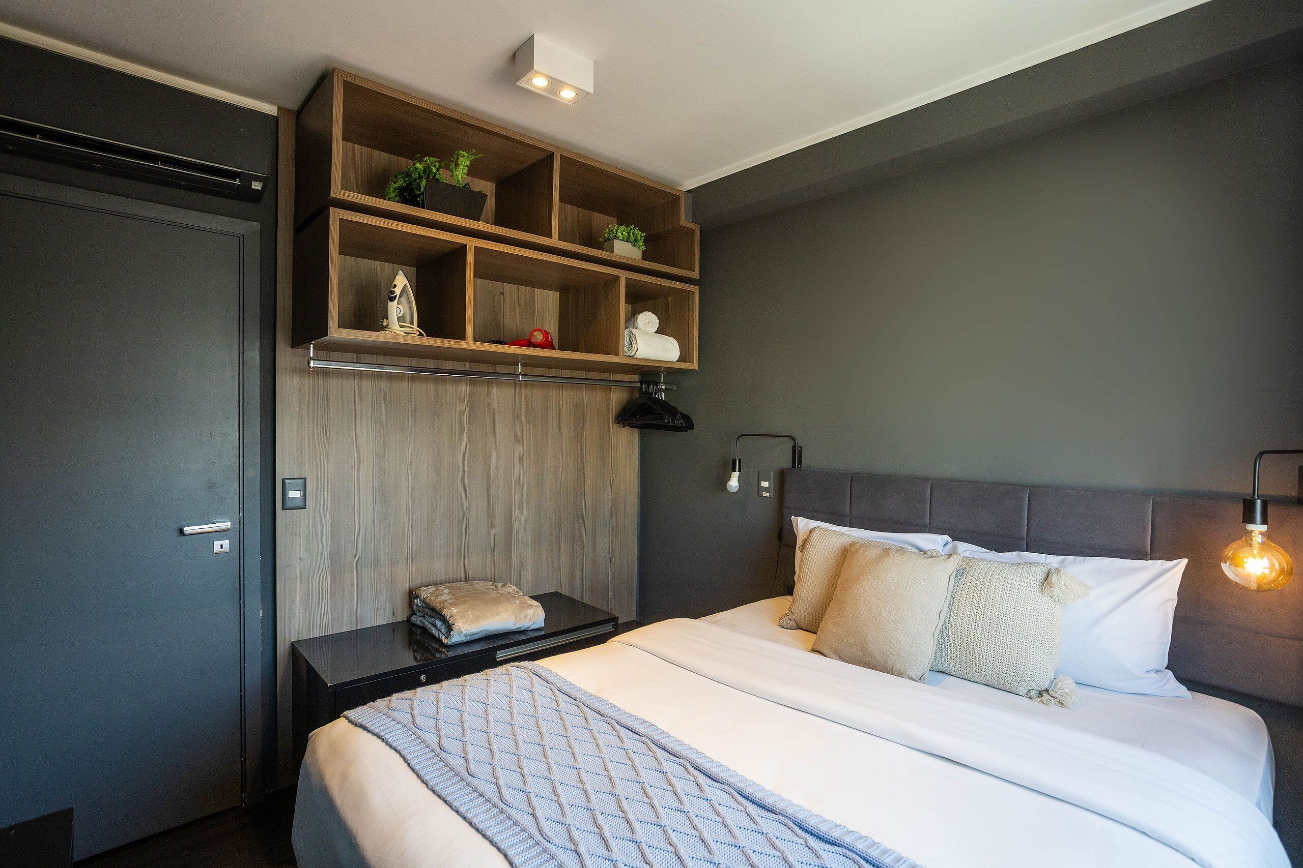 A modern hotel room with a bed, a nightstand, a wooden shelf with plants, a wall-mounted light, and a door.