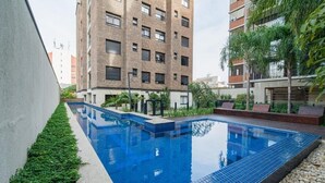 Una piscina con borde de azulejos, rodeada de follaje y contigua a un edificio de varios pisos.