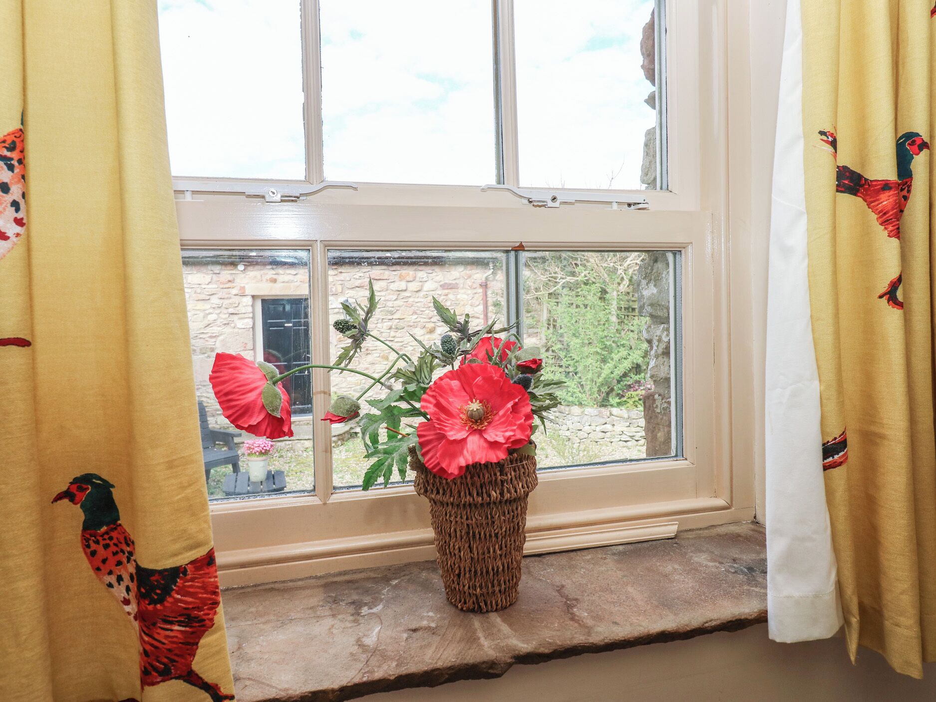 A window with a woven basket containing red flowers, a stone wall, and a small garden visible through the window.