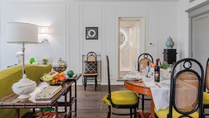A dining area with a table set for four, surrounded by wicker chairs with yellow cushions. A lamp with a white shade is on a side table, and a bowl of fruit is on the table. - Casa Antonia in Lucca (Lucca)