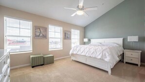 Une chambre avec un grand lit, un ventilateur de plafond et deux fenêtres équipées de stores.