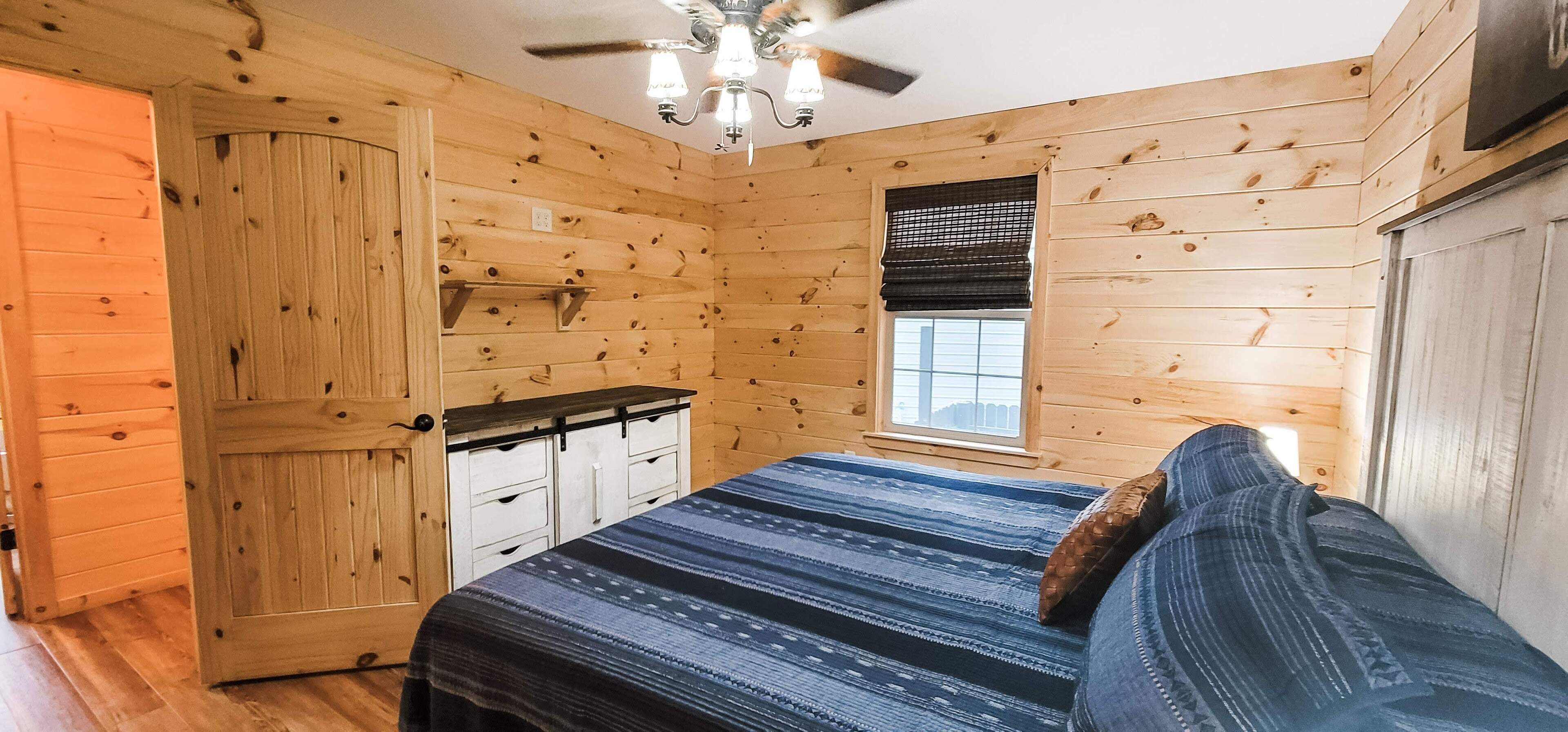 A wooden cabin bedroom with a bed, a ceiling fan, a window with blinds, and a door.