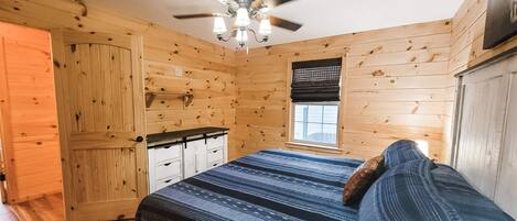 Un dormitorio de cabaña de madera con una cama, un ventilador de techo, una ventana con persianas y una puerta.